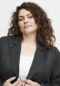 Garnitur w kolorze węgla o dopasowanym kroju, z wykładanymi klapami i subtelną teksturą. Nosi się go na białej bluzce wykończonej koronką. Ręce umieszczone na brzegach marynarki.