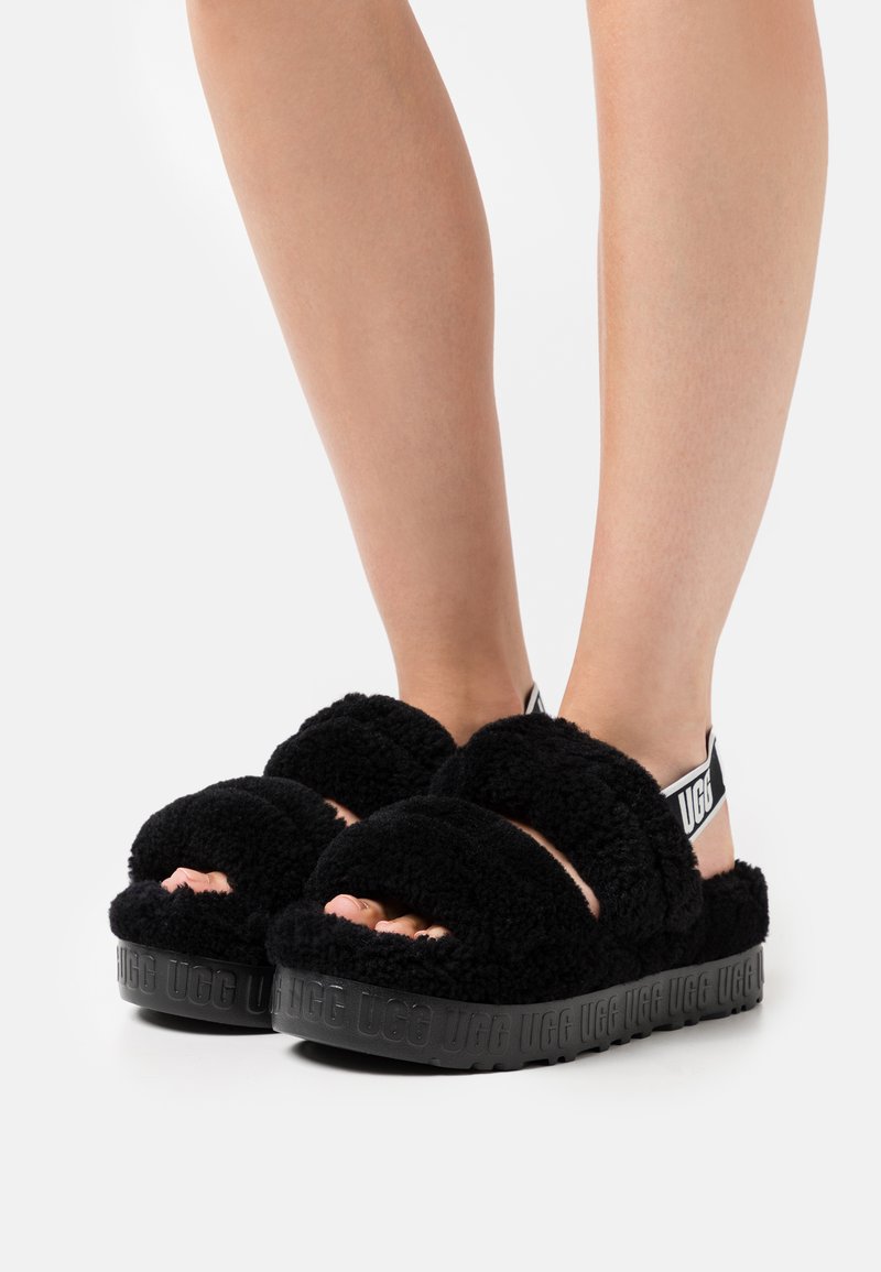 Sandalias de slide negras y esponjosas con tiras anchas, punta abierta y suela de goma con el logo "UGG" grabado. Material superior suave y texturizado.