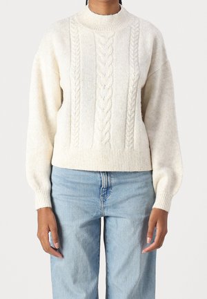 Person iført en cremefarvet kabelstrikket sweater med lange ærmer og høj hals, kombineret med lyseblå denimjeans.