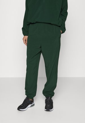 Persoon die donkergroene losse broek en bijpassende top met lange mouwen draagt, met zwarte en witte Nike-sneakers, staand op een effen vloer.