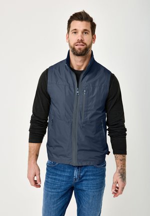 Man with beard and tattoo wearing navy sleeveless zip-up vest over black long-sleeve shirt and blue jeans, standing against light background.
