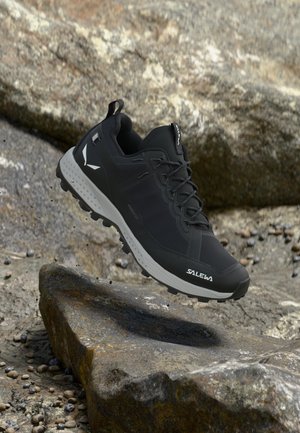 Μαύρο και γκρι ορειβατικό παπούτσι Salewa με ανθεκτική σόλα, αιωρούμενο πάνω από βραχώδες έδαφος.