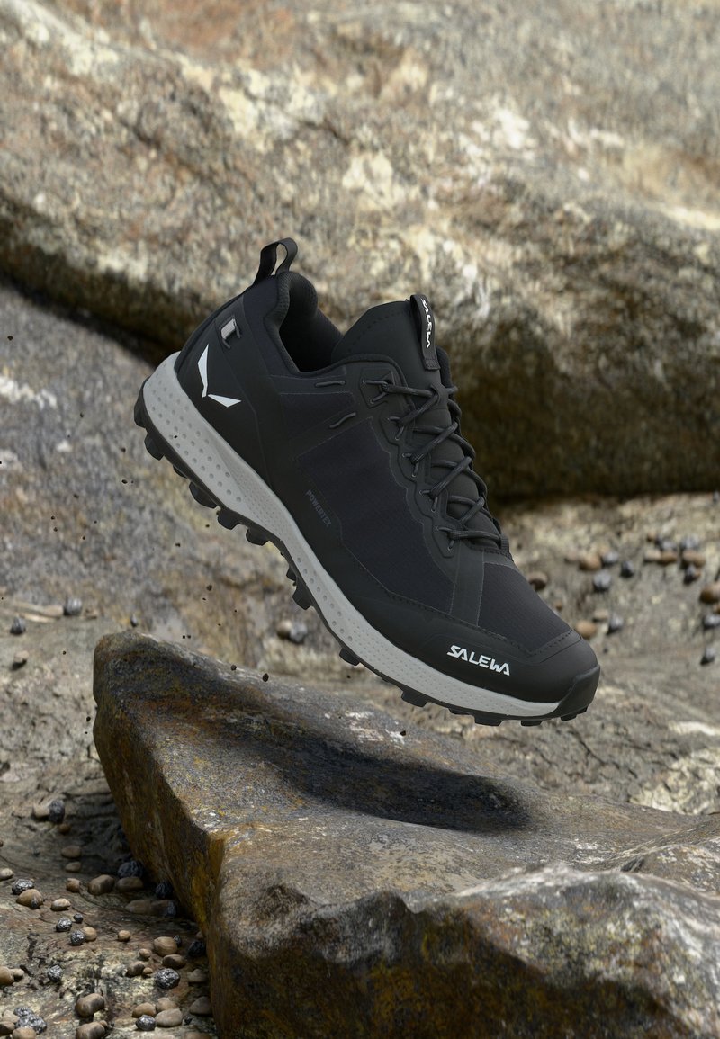 Μαύρο και γκρι ορειβατικό παπούτσι Salewa με ανθεκτική σόλα, αιωρούμενο πάνω από βραχώδες έδαφος.