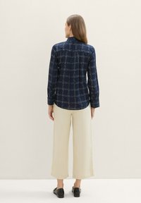 Chemise bleu marine à boutons, avec un motif à carreaux blancs, manches longues et col ; associée à un pantalon large beige et des chaussures noires.