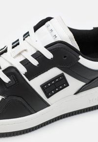 Sneaker in ecopelle bianca e nera con lacci bianchi, dotata di suola in gomma e logo "TOMMY JEANS" sul lato.