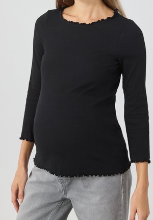 Frau trägt ein eng anliegendes, schwarz geripptes Langarmshirt mit gewelltem Saum und hellgraue Jeans, steht vor einem neutralen Hintergrund.