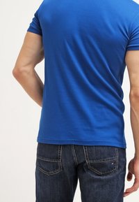 Niebieska bawełniana koszulka z krótkim rękawem, dopasowany fason, gładka faktura. Noszona z ciemnymi jeansami z tylnymi kieszeniami i ozdobnymi szwami.