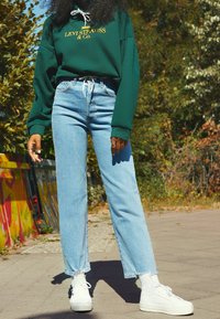 Sudadera verde con logo amarillo, jeans de cintura alta azules y zapatillas de plataforma blancas. El atuendo es casual y de corte relajado.