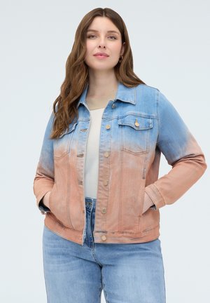 Junge Frau trägt eine blau- und pfirsichfarbene Ombre-Jeansjacke über einem weißen Top und hellblauen Jeans, steht mit den Händen in den Taschen.