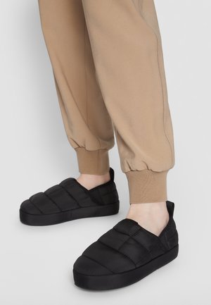 Fødder iført sorte quiltede slip-on sko parret med beige bukser med opbuk mod en enkel lys baggrund.
