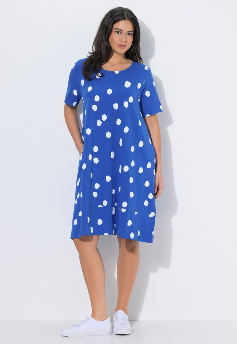 Frau steht in einem knieumspielenden blauen Kleid mit weißen Polka-Dots und weißen Sneakern vor einem schlichten hellen Hintergrund.