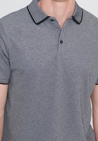 Hombre que lleva una camiseta polo gris con cuello y bordes de las mangas a rayas blancas y negras, parcialmente abotonada en el cuello.