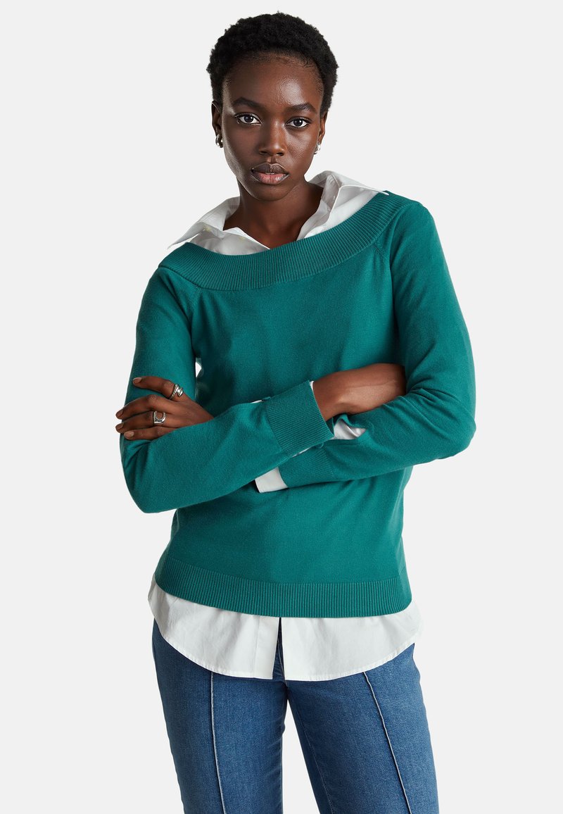 Modrý svetr s žebrovaným výstřihem a manžetami, vrstvený přes bílou košili s límečkem. Má přiléhavý střih a měkkou texturu.