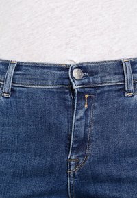 Jeans de denim de color azul oscuro, con un botón plateado y costura amarilla. El detalle del bolsillo deshilachado añade textura a la tela.