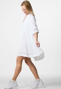 Weißes, knielanges Kleid mit kurzen Ärmeln, strukturiertem Stoff, gestuftem Rüschensaum und Spitzenverzierungen. Kombiniert mit weißen Turnschuhen.