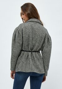 Grå og hvid jakke med houndstooth-mønster, krave, puffede ærmer og et sort bælte i taljen, lavet af tekstureret stof.