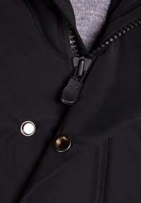 Zbliżenie na zamek błyskawiczny czarnej kurtki częściowo otwarty na szarym swetrze dzianinowym, z dwoma metalowymi guzikami zatrzaskowymi widocznymi na tkaninie.