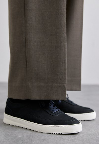 Black suede sneakers with white rubber soles, featuring a rounded toe and laces, worn with gray tailored pants.