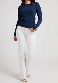 Marineblaues, gestricktes Langarmoberteil mit figurbetontem Design und dezentem Branding, kombiniert mit weißen, ausgestellten Jeans und weißen Sandalen mit Riemchen.