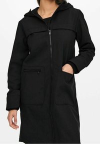 Una mujer que lleva un abrigo negro con capucha y cremallera frontal, posando con una mano en un bolsillo, mostrando el diseño y el ajuste del abrigo.
