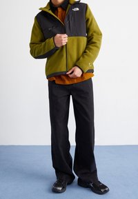 Groen-zwart fleecejack met een rits aan de voorkant, twee borstzakken, een ronde kraag, gedragen over een oranje shirt, gecombineerd met zwarte broek.