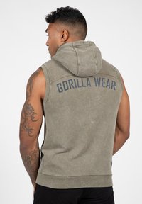 Mouwloze hoodie in olijfgroene stof met een textuurafwerking. Heeft een kap en het "GORILLA WEAR" logo op de achterkant gedrukt.