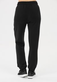 Zwarte joggingsbroek van zachte stof met een elastische tailleband en enkelmanchetten. Draagt zwart sport schoenen met witte accenten.