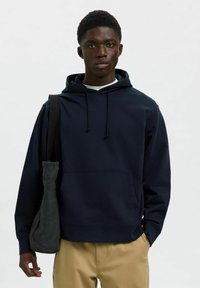 Sudadera con capucha azul marino, con cordones, bolsillo frontal y textura suave. Combinada con pantalones beiges y un bolso tote gris.