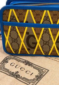 Sac Gucci marron et jaune à motif avec une bordure bleue reposant sur un sac anti-poussière beige Gucci avec une bordure noire décorative et un texte.