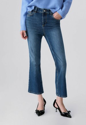 Person trägt blaue, kurze Schlagjeans kombiniert mit schwarzen spitzen High Heels mit Schleifendetails und einen hellblauen Pullover.