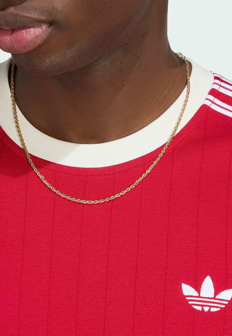 Gros plan sur une personne portant un t-shirt rouge Adidas avec un col blanc et des rayures, ainsi qu'un collier chaîne torsadée dorée autour du cou.
