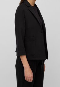 Blazer negro con un diseño estructurado, fabricado en tejido texturizado. Presenta un cuello con muesca, dos bolsillos frontales y mangas de tres cuartos.