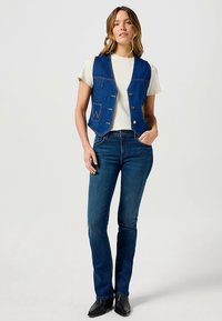 Tmavomodrá rifľová vesta s priliehavým strihom, s tromi gombíkmi a predným vreckom. Nosená cez svetlú tričko a modré džínsy.