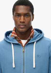 Felpa con zip di colore azzurro chiaro, con lacci bianchi e zip in ottone, indossata sopra una camicia a quadri nei toni dell'arancio, del verde e del blu.