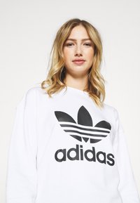 Bijela dukserica s velikim crnim Adidas logotipom i tri pruge te trefoil dizajnom sprijeda. Mekana tekstura, opušteni kroj.