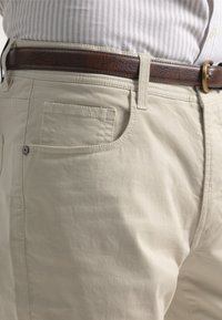 Pantalones de algodón beige con una textura suave, que cuentan con un bolsillo frontal y un cinturón de cuero marrón con una hebilla dorada. Camisa a rayas visible arriba.