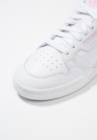 Sneaker in pelle bianca con punta perforata, lacci bianchi piatti e sottili accenti rosa sul lato. Suola arrotondata. Finitura testurizzata.