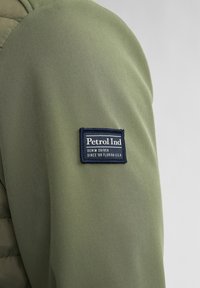 Giacca di tessuto verde oliva con spalla, dotata di una toppa rettangolare blu navy etichettata "Petrol Ind", con testo bianco e dettagli ricamati.
