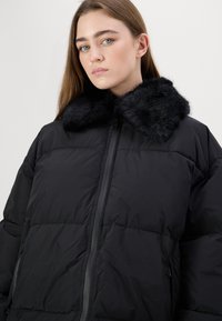 Black puffer jacket with a faux fur collar. Features a zip front, quilted texture, and wide-cut sleeves. Smooth material and structured design.