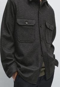 Camicia di flanella grigio scuro con due tasche sul petto con bottone, orlo arrotondato e texture morbida. Presenta bottoni neri e un design con colletto aperto.
