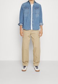 Denimskjorta i ljusblått med två bröstfickor, knappstängning och uppvikta ärmar; kombinerad med beiga byxor och vita sneakers med marinblå snören.