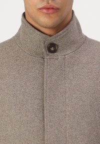 Brun ullblandingsfrakk med høy krage, med synlig knappeåpning og strukturert stoff. Enkel design, uten pynt.
