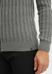 Grauer Kabelstrickpullover mit gerippten Bündchen und Saum. Verfügt über ein strukturiertes Muster und ein kleines braunes Logo-Label an der unteren linken Naht.