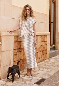 Femme en haut rayé beige et jupe, appuyée contre un mur en pierre, souriante, avec un petit chien noir marchant à proximité dans une rue pavée.