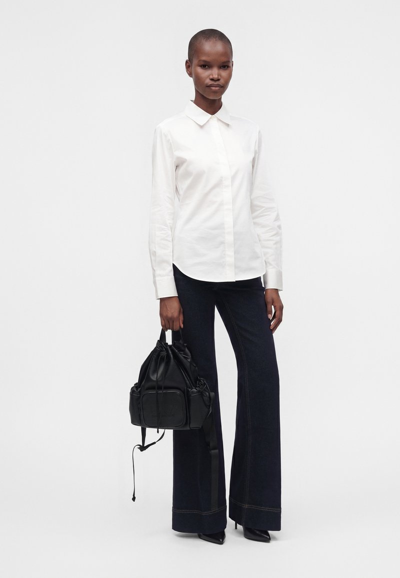 Femme en chemise blanche à manches longues et pantalon large foncé tenant un sac à main noir sur fond blanc uni.