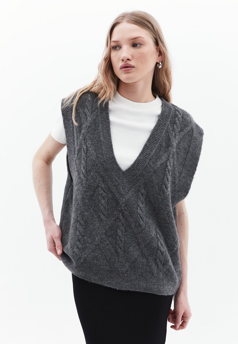 Gilet grigio lavorato a maglia con scollo a V, caratterizzato da un motivo a trecce e maniche corte, indossato sopra una camicia bianca, su uno sfondo chiaro.