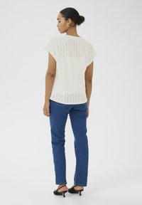 Haut blanc à manches courtes avec un design perforé ; associé à un jean droit bleu et des sandales à talons noires ; vu de l'arrière.