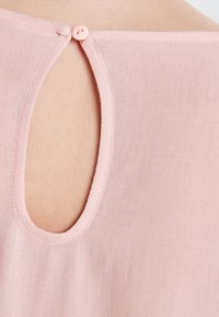 Světle růžový top z látky s kulatým výřezem na zádech, zajištěný malým odpovídajícím knoflíkem, s hladkou strukturou a jednoduchým šitím.