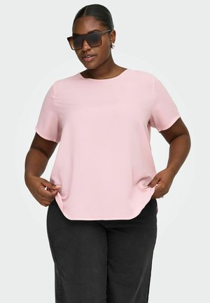 Kvinde med mørk hud iført store, mørke solbriller, lyserød kortærmet bluse og sorte bukser, der blidt holder i blusens sider.