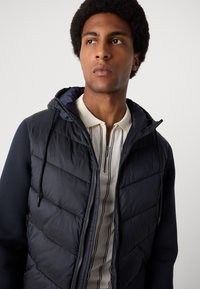 Navy puffer jacket with a hood, featuring quilted texture and zippered front. Paired with a light-colored striped polo shirt underneath.